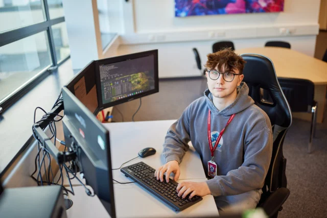 A young man sits at a desk with two computer monitors, focused on work.
