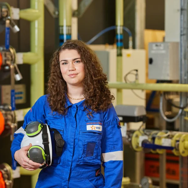 A woman in a blue uniform holding a safety helmet in an industrial setting.