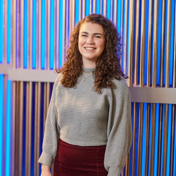 A young woman with curly hair smiling in front of a blue striped background.