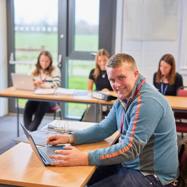 Young man sitting at a desk with a laptop in a classroom setting.
