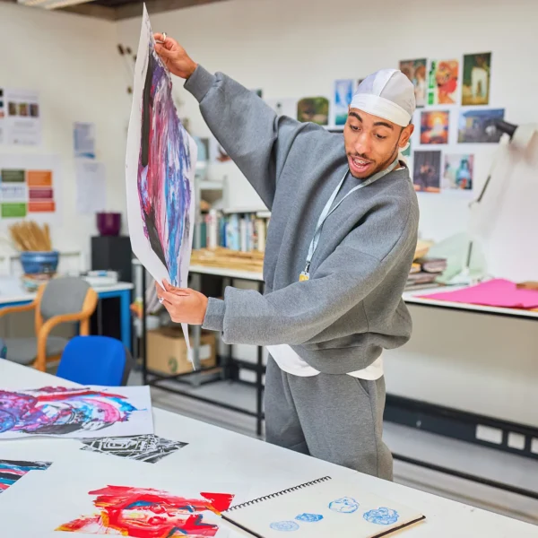 A man in a grey tracksuit displaying a piece of art in a classroom setting.