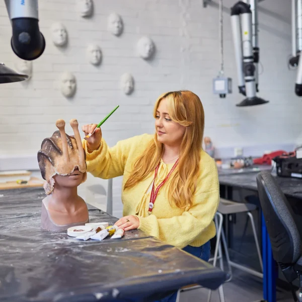 A female makeup artistry student in a yellow top painting a human head mannequin shaped like a snail shell with a green brush.