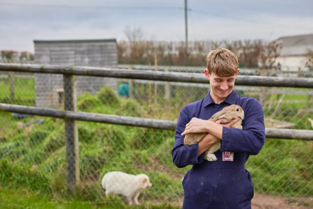 a young man holding a rabbit outside a fenced area