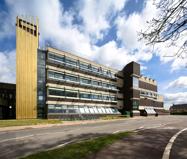 A modern multi-storey building with a tall tower on the left and a road in front.