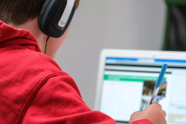 A child wearing headphones uses a laptop and writes with a pen.