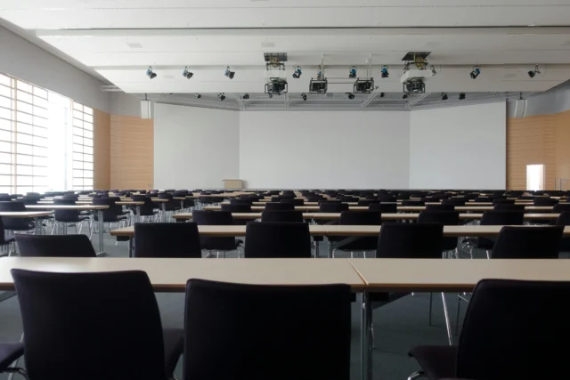 A large empty classroom with rows of desks and chairs, and a projector screen on the wall.