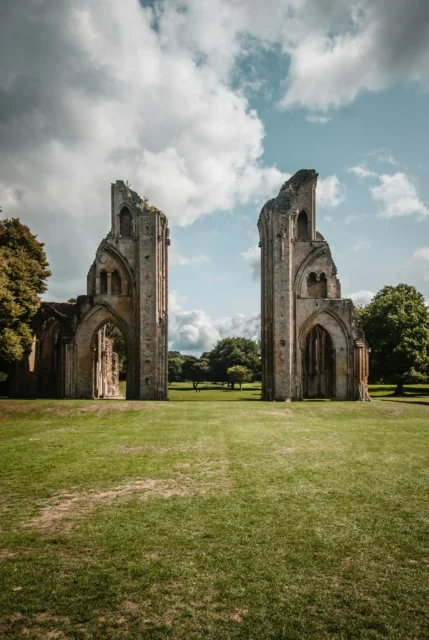 Two tall, ruined towers stand on green grass with trees and blue sky in background.