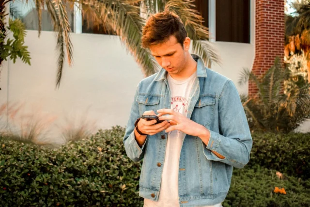 Young man in denim jacket looking at his phone outdoors.