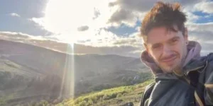 Man smiling in a scenic landscape with a bright sun.