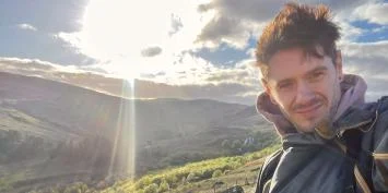 Man smiling in a scenic landscape with a bright sun.