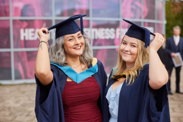 Two women in graduation gowns and caps, smiling and posing for a photo.