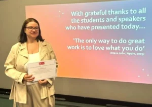 Krystyna stands holding a certificate in front of a presentation slide at the UWE Bristol conference with a thank-you message and an inspirational quote by Steve Jobs.