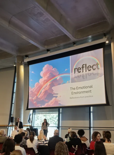 Audience watching a presentation titled 'Reflect: The Emotional Environment' at a conference or seminar, with a vibrant slide featuring pink clouds and a rainbow displayed on the screen.
