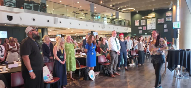 A group of people gathered in a spacious venue, attentively participating in an event or presentation led by a speaker.