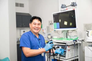 A smiling medical professional in blue scrubs and gloves holds an endoscope in a modern diagnostic room.