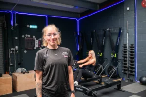 Personal trainer standing confidently in front of modern gym equipment, with another client working out in the background.