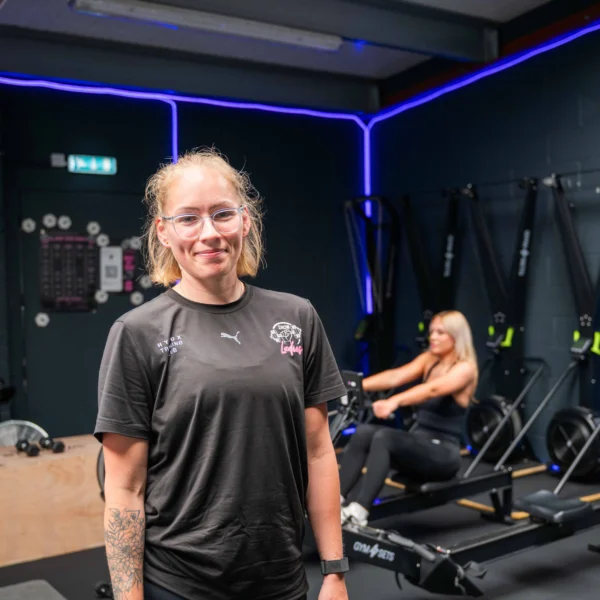 Personal trainer standing confidently in front of modern gym equipment, with another client working out in the background.