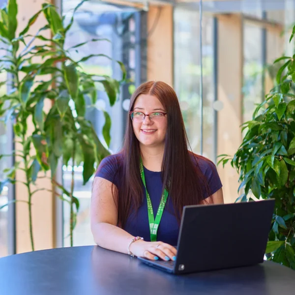 A woman with long brown hair and glasses is sitting at a table in a bright, plant-filled room, working on a laptop.