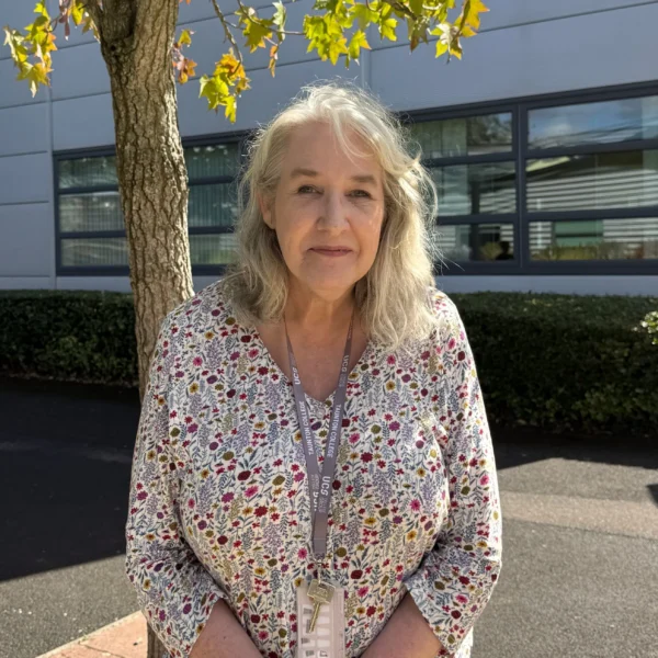 Pam Jones, a mature woman with curly grey hair, wearing a floral blouse, seated outdoors in front of a modern office building.