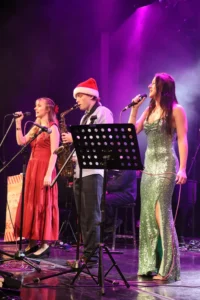 Three musicians performing on stage, a man with a Santa hat and two women in evening gowns.
