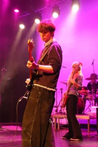 Man playing guitar on stage with a drummer in the background.