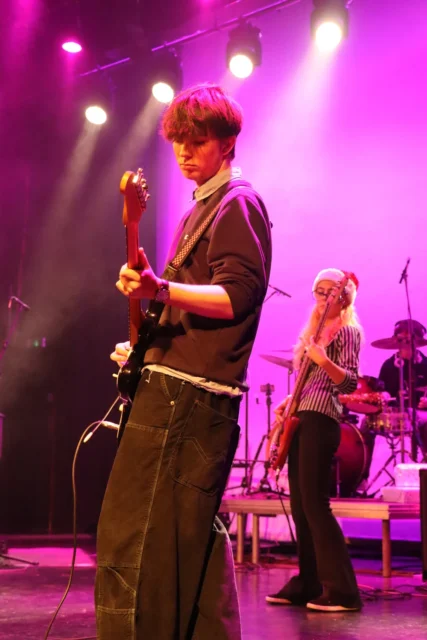 Man playing guitar on stage with a drummer in the background.
