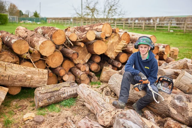 A smiling child in safety gear sitting on logs with a chainsaw.