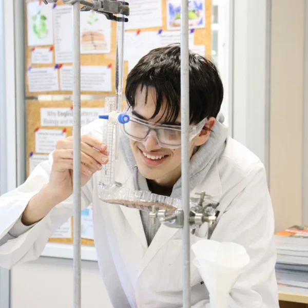A young Asian man in a white lab coat and safety glasses holds a test tube and rack in a laboratory setting.