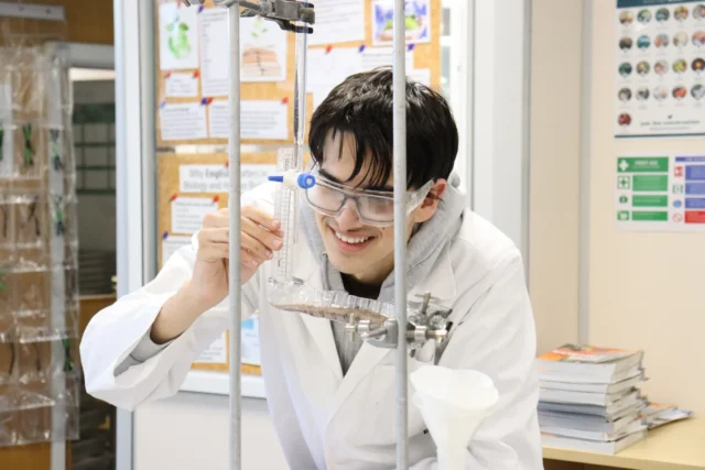 A young Asian man in a white lab coat and safety glasses holds a test tube and rack in a laboratory setting.