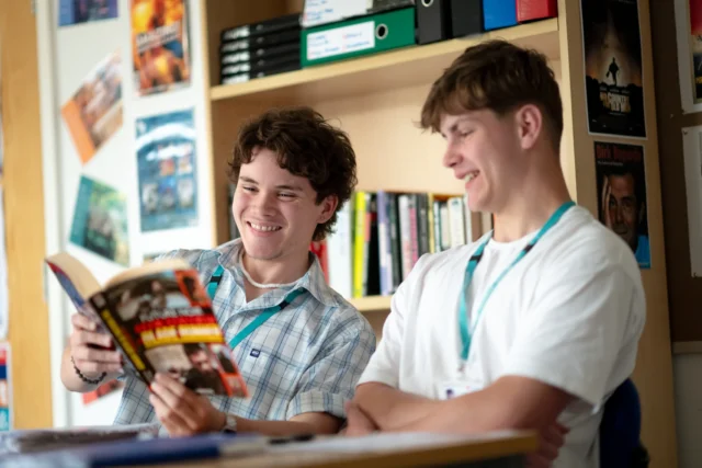 Two teenage boys in a classroom setting looking at a book together.