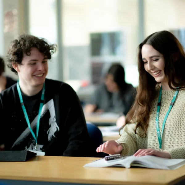 A young man and woman sitting at a desk in a classroom, both wearing lanyards and engaged in learning.