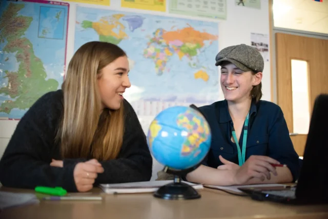 Two young women sitting at a desk with a globe looking at each other.