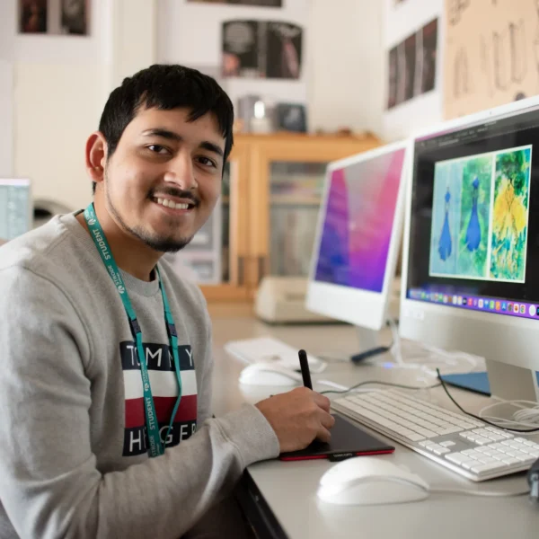 A young man sitting at a computer with a colorful image on the screen, smiling and looking at the camera.