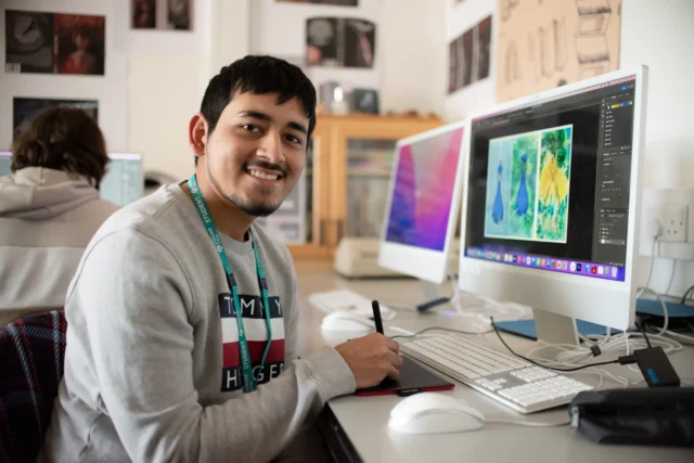 A young man sitting at a computer with a colorful image on the screen, smiling and looking at the camera.