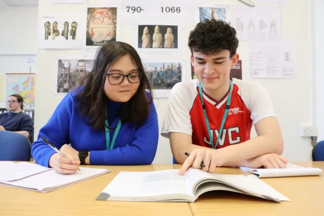 Two students sitting at a table with an open book and papers, engaged in studying or working on a project.