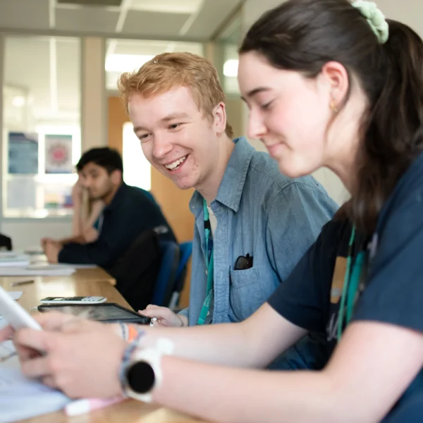 Two students sitting at a desk, looking at papers and smiling. They appear to be in a classroom or study area.