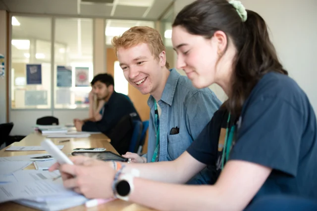 Two students sitting at a desk, looking at papers and smiling. They appear to be in a classroom or study area.