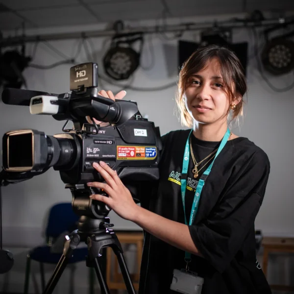 Woman operating a large video camera, smiling and wearing a lanyard around her neck.