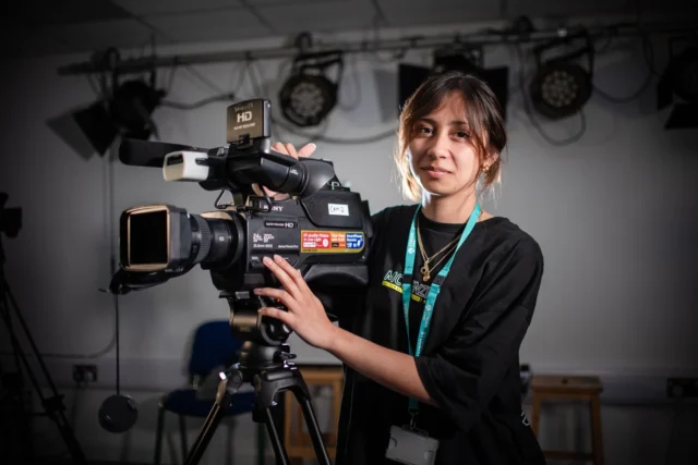 Woman operating a large video camera, smiling and wearing a lanyard around her neck.