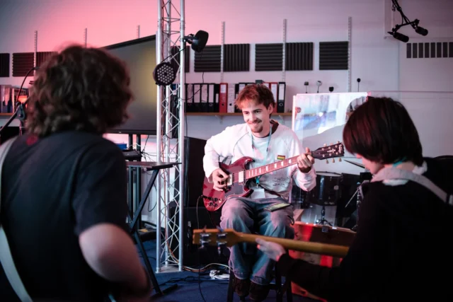 A man playing an electric guitar while two people watch, likely in a music classroom or rehearsal setting.