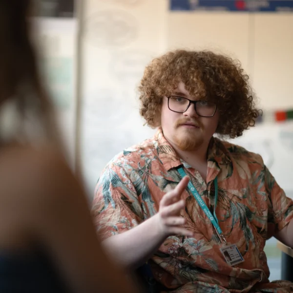 A person with curly hair and glasses gesturing while talking to another person.