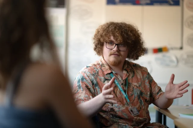 A person with curly hair and glasses gesturing while talking to another person.