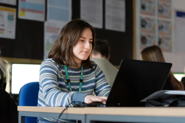 A young girl with brown hair sitting at a desk with a laptop on it.
