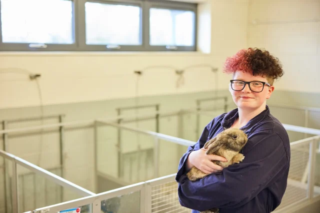 A woman with short hair and glasses holding a dog, smiling at the camera in an animal shelter.