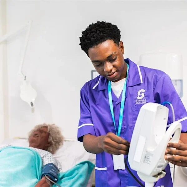A male nurse in purple scrubs examining a medical device beside a patient.