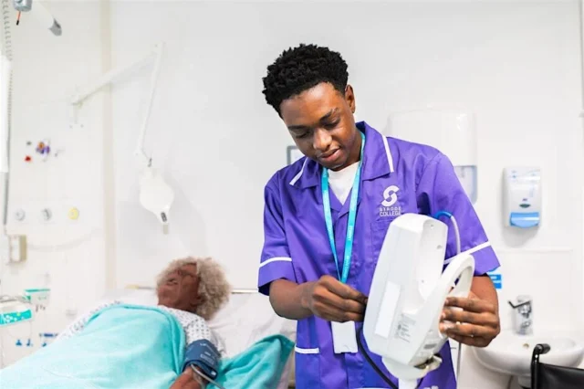 A male nurse in purple scrubs examining a medical device beside a patient.