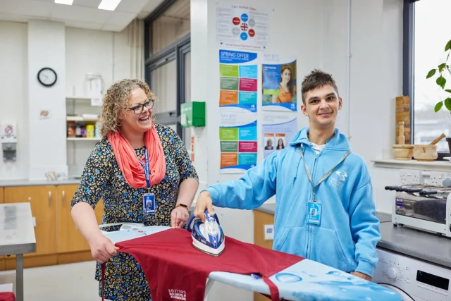 A woman and a boy ironing a red sheet in a classroom setting.