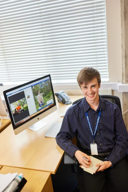 A young man sitting at a desk with a computer monitor displaying an image.