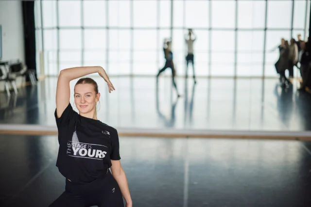 Young dancer in a studio with large windows, performing a pose.
