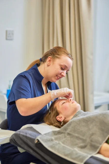 A woman lies on a treatment bed while a technician examines her face.